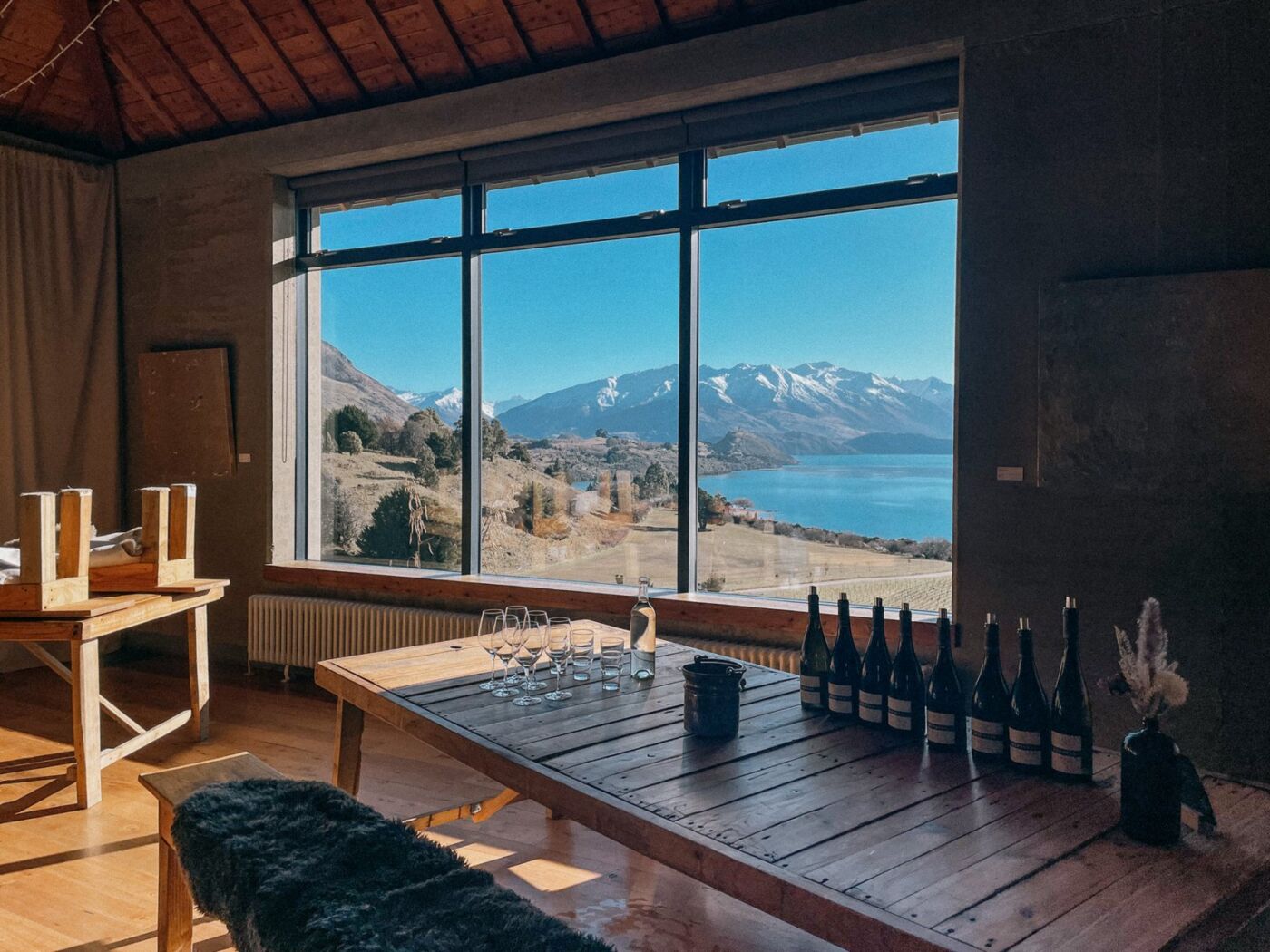 Inside the Rippon cellar door, a long timber table set with glasses and a row of wine bottles beneath large windows looking out over the vineyard, Lake Wānaka and the mountains