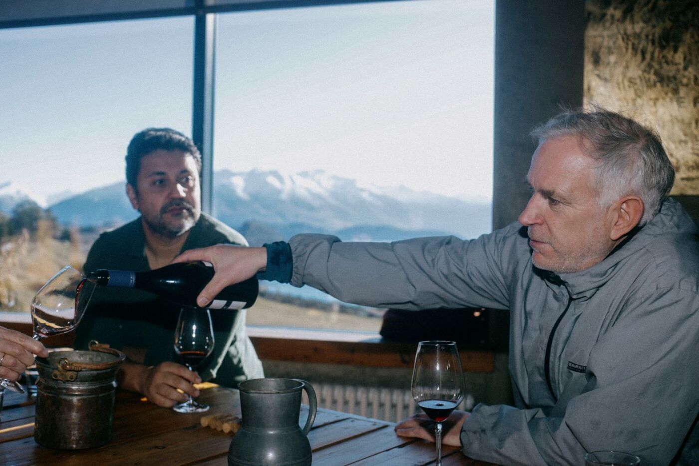 Nick Mills reaching across the tasting table to pour red wine into a tilted glass, with Lake Wānaka visible through the window behind