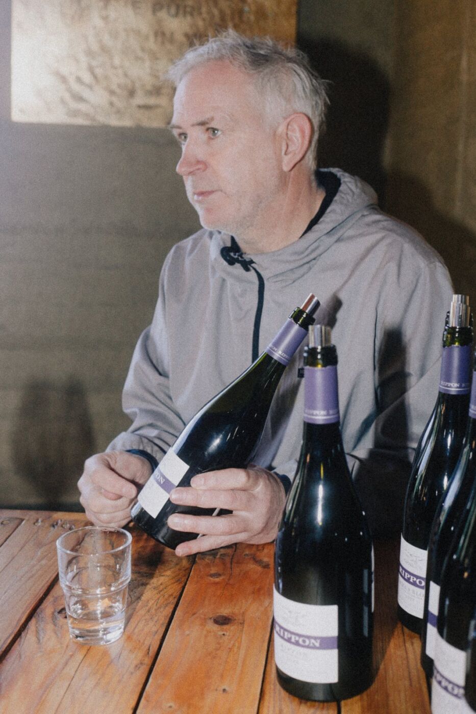 Nick Mills seated at the timber tasting table holding a bottle of Rippon wine, with more bottles wearing the purple-banded label in the foreground