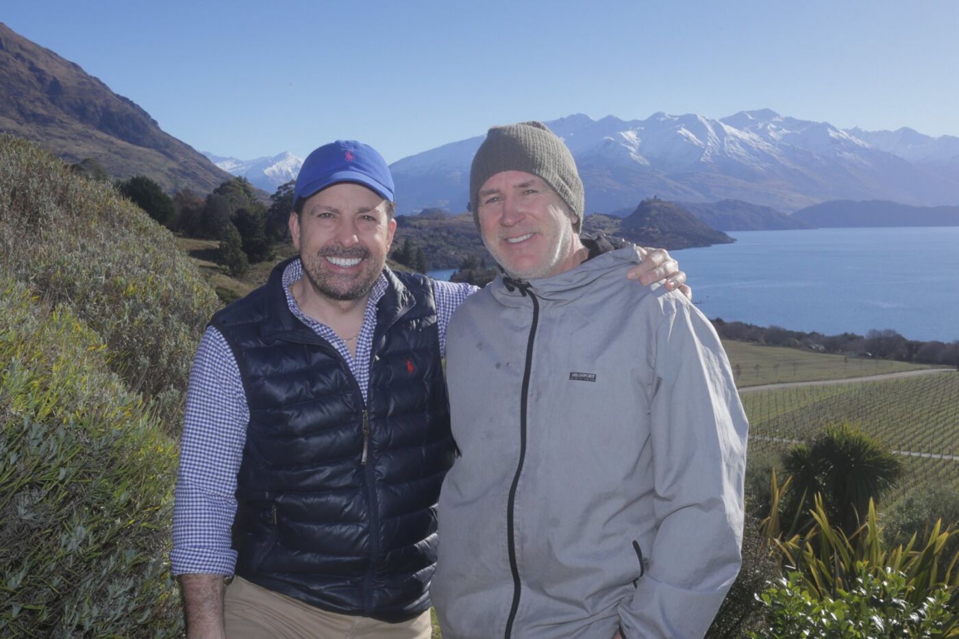 Two men smiling in front of the Rippon vineyard, with vine rows, Lake Wānaka and snow-covered mountains in the background