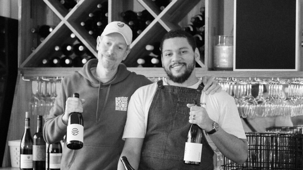 Black and white photo of two people holding bottles of wine in front of the wine rack at Bar Septimus