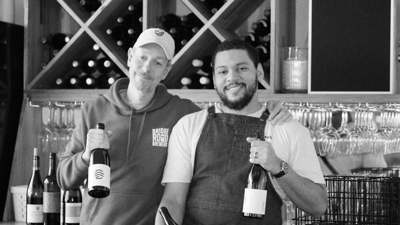 Black and white photo of two people holding bottles of wine in front of the wine rack at Bar Septimus