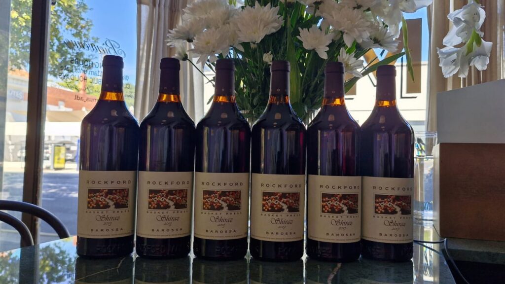 A row of six bottles of Barossa Shiraz lined up in the front window at Bar Septimus with white flowers behind